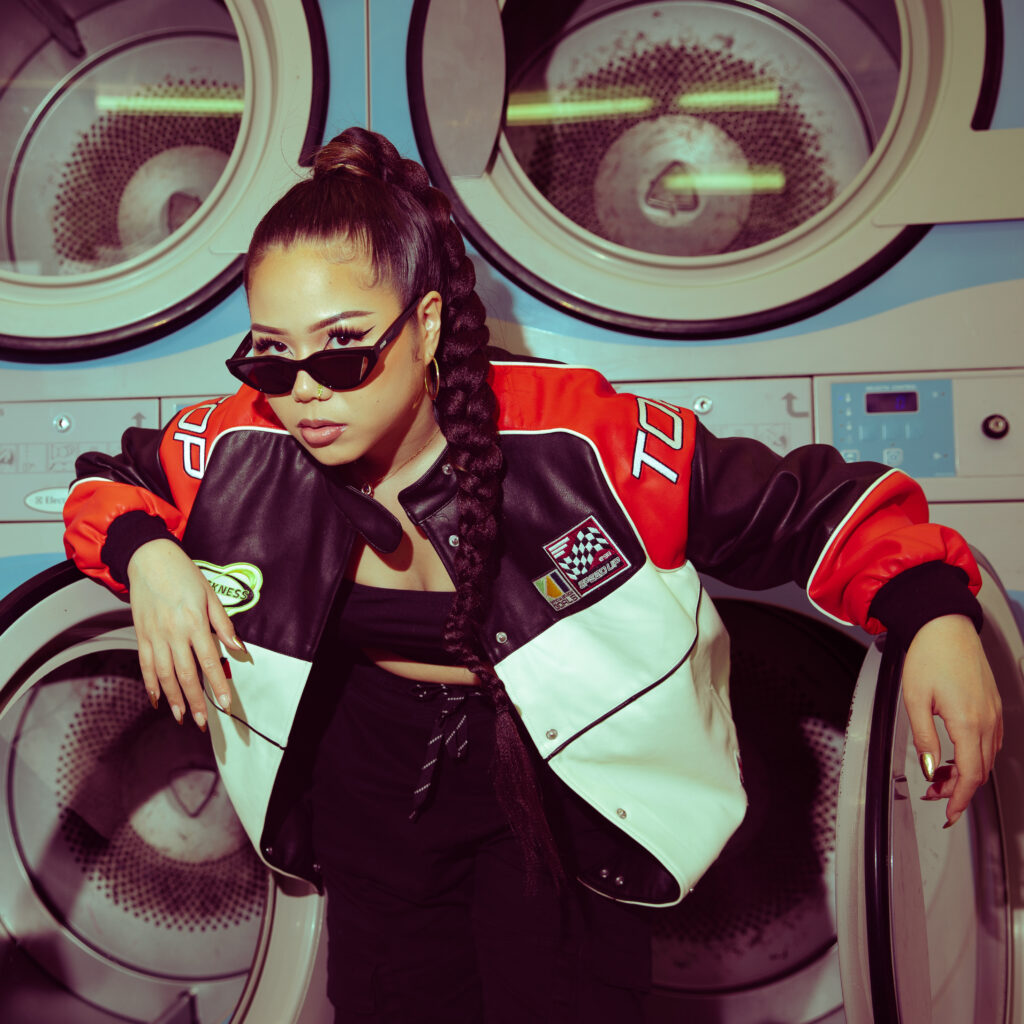 Photo Femme prise en plan large devant une machine à laver dans un Lavomatic, veste de motard, lunettes noire larges, lumière au flash, tresse longue, couleur vintage style années 90. Photographe professionnel, direction artistique
