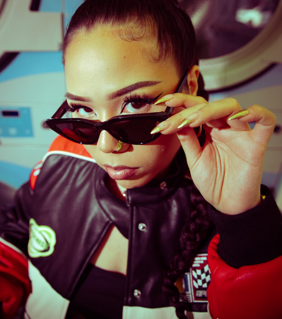 Photo Femme prise en plongée devant une machine à laver dans un Lavomatic, veste de motard, lunettes noire larges, lumière au flash, tresse longue, couleur vintage style années 90