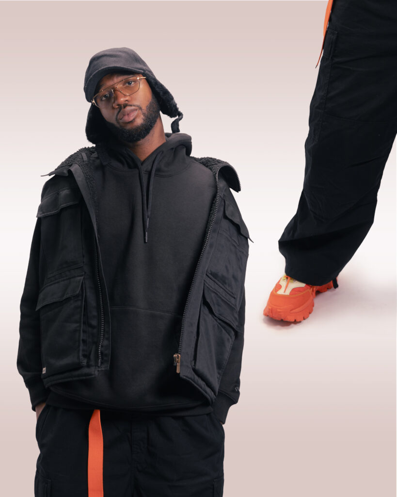 shooting en studio, homme noir habillé avec des vêtement sombre. Veste entrouverte noire gillée sur les épaules. Photomontage avec ajout à droite de la jambe du modèle. Arrière plan beige clair. Photographe professionnel, direction artistique.